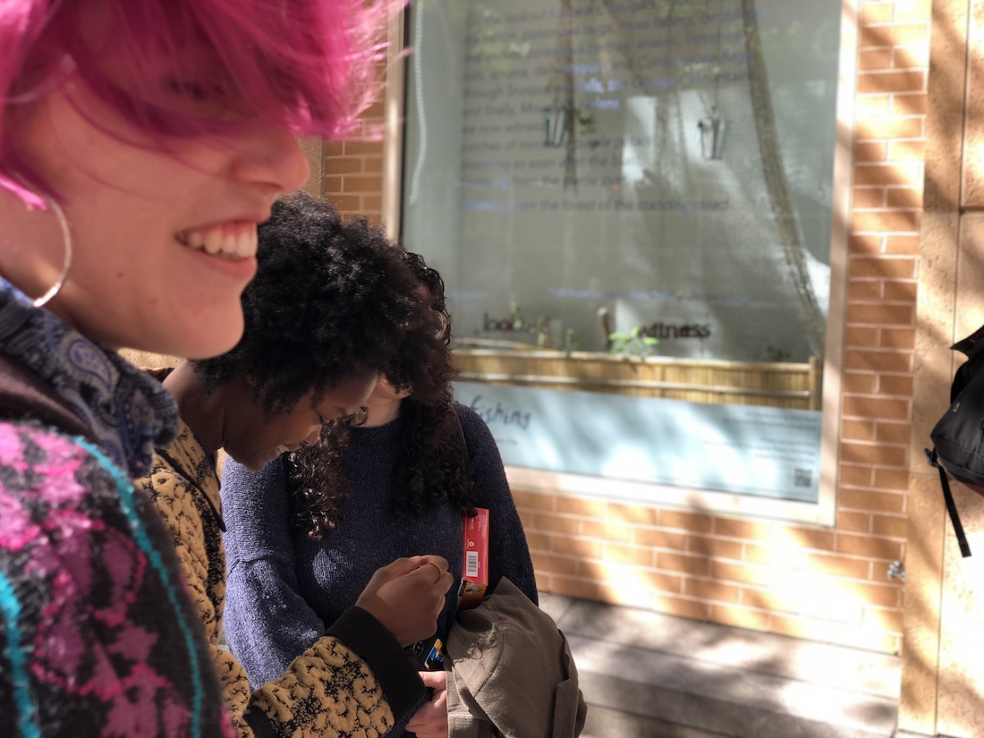 the side of a woman's face with pink hair and a smile in foreground, behind her is a black woman looking down and smiling